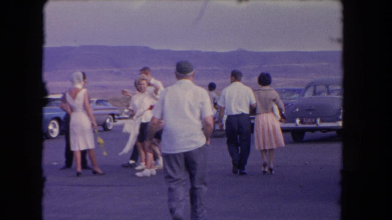 1962 Los Angeles-area home-movie frame showing adults in summer clothing walking between parked classic cars near distant hills.