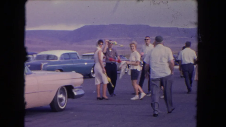 Adults in 1960s clothing stand and walk near a pink car, blue sedan, and other parked vehicles in a Los Angeles-area lot.