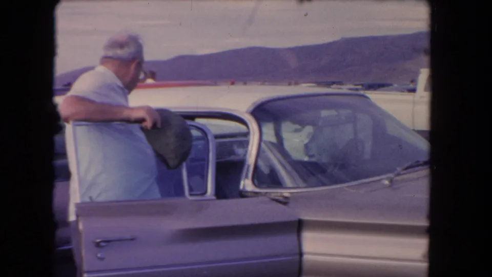 A man in a light shirt stands at an open classic car door, with more vehicles and hazy hills visible in the background.