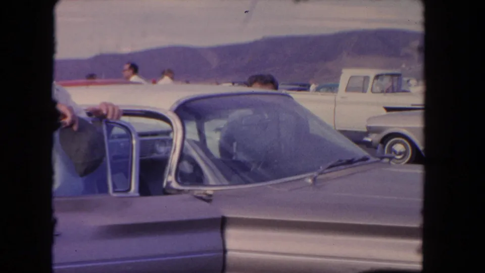 Close 8mm frame of a classic car with an open door, partial figures, a white pickup, and hills beyond the parking area.