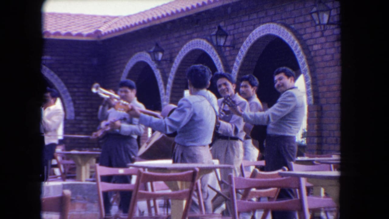 In a 1962 Los Angeles home-movie frame, mariachi musicians play in a brick courtyard beneath arched colonnades.