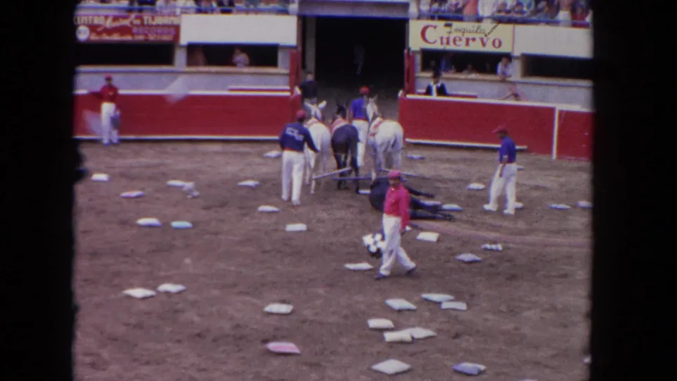 1962 Los Angeles home-movie frame showing a flag carrier crossing the dirt arena as mounted handlers and attendants remain gathered around the bull.