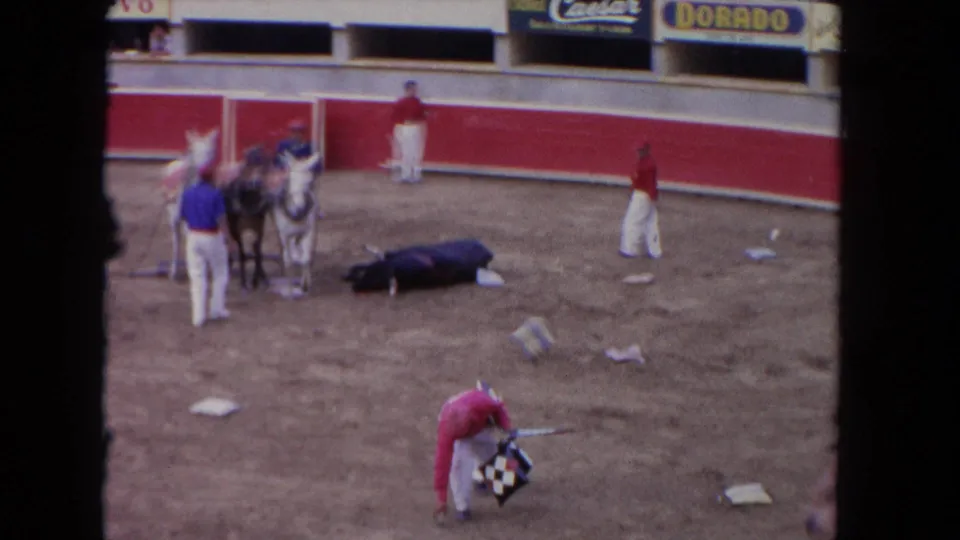 1962 Los Angeles arena frame showing a bullfighter bending near a patterned cloth, with horses, attendants, and a fallen bull behind him.