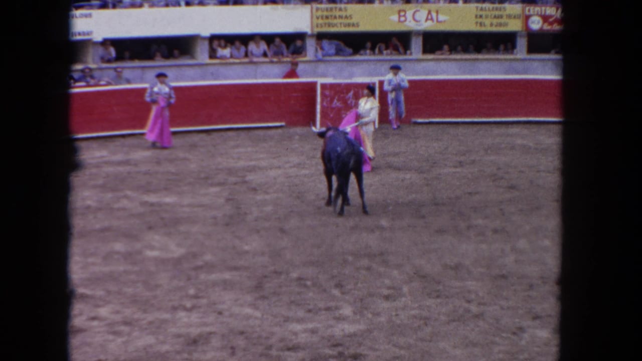 1962 Los Angeles home-movie frame showing a wide bullring view with a matador, a black bull, red arena boards, spectators, and signage.