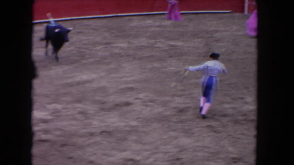 1962 Los Angeles home-movie frame of a matador moving away from a black bull inside a red-walled arena.