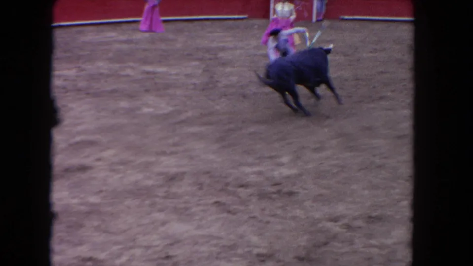 1962 Los Angeles 8mm frame showing a black bull turning near a performer with a pink cape on the dirt arena floor.