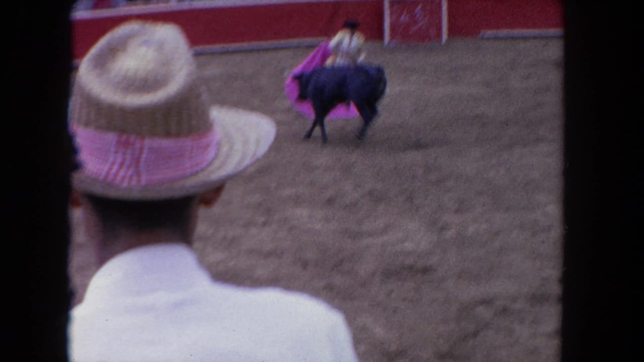 1962 Los Angeles home-movie frame showing a straw-hatted spectator in the foreground as a man works a pink cape beside a dark bull in an arena.