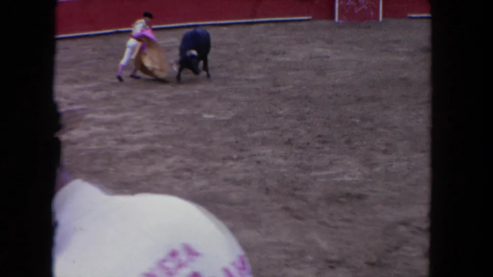 1962 Los Angeles home-movie still showing the caped figure close to the bull in the sandy ring, framed partly by a foreground spectator and film borders.