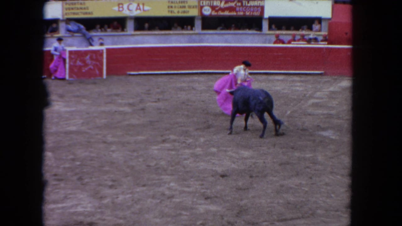 1962 Los Angeles 8mm frame with a bull moving toward a matador's pink cape near red arena barriers and spectators.