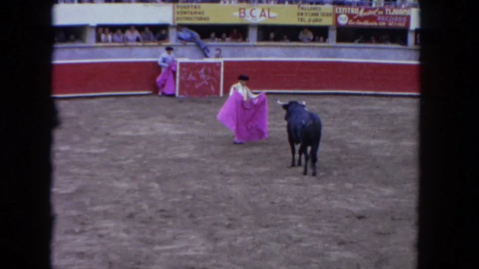 1962 Los Angeles 8mm frame of a matador in a light suit and black hat holding a pink cape before a black bull.
