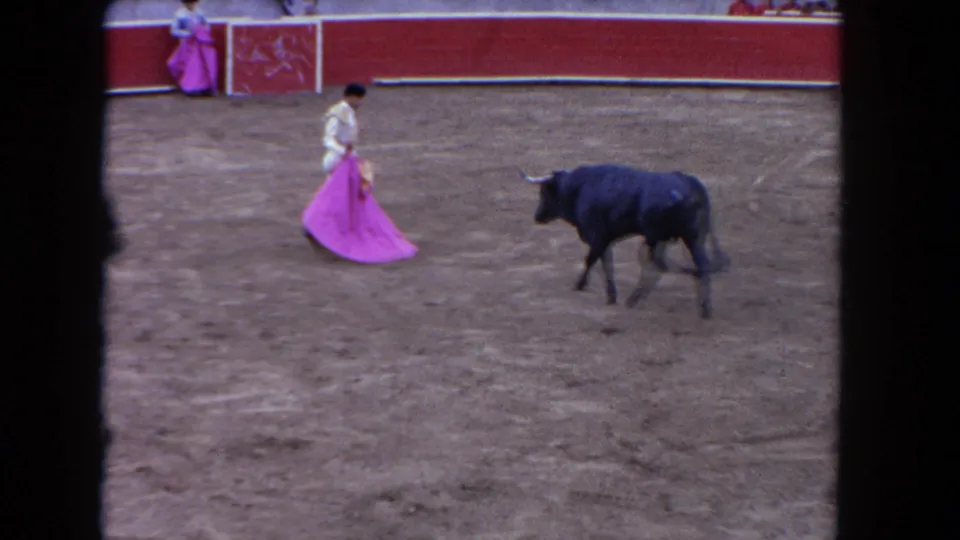 1962 Los Angeles arena frame with a matador holding a pink cape as a black bull stands across the dirt ring near red-and-white barriers.