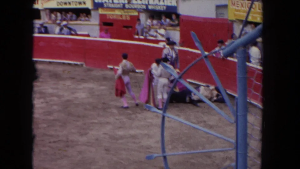 1962 Los Angeles arena view with matadors carrying capes near a bull, spectators in the stands, and painted signs above the red wall.