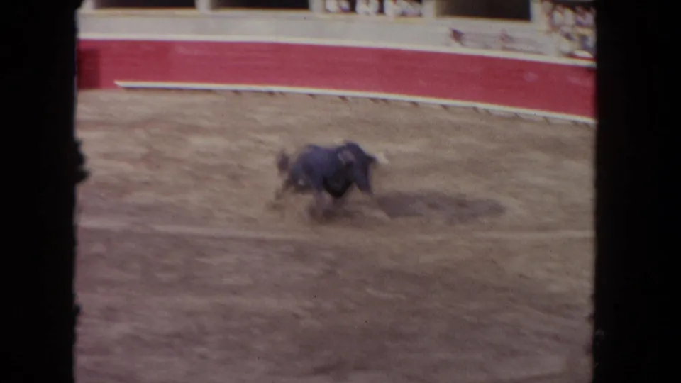1962 Los Angeles 8mm frame showing a bull running or turning in the dirt ring, with red arena wall and spectators beyond.