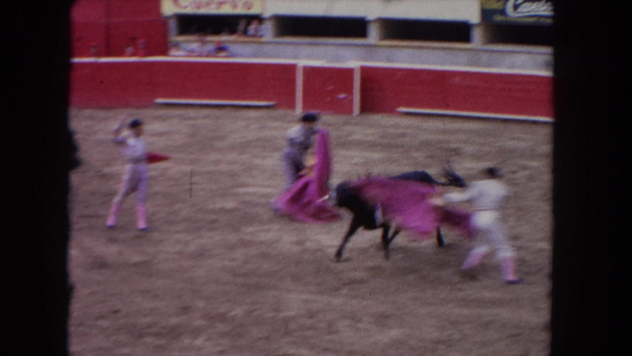 1962 Los Angeles home-movie frame showing matadors in pale suits maneuvering pink capes around a charging black bull in an arena.
