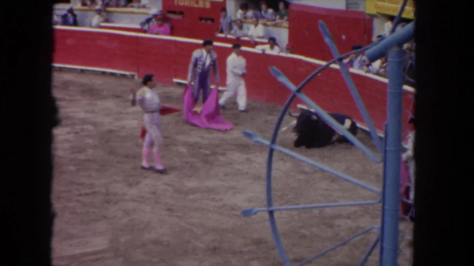 Home-movie frame from the Los Angeles bullring sequence, with matadors in pale and patterned suits near a black bull and visible spectators.