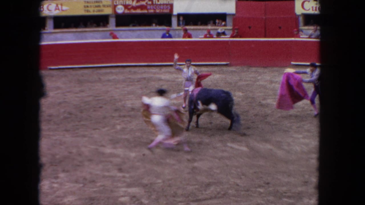 1962 Los Angeles 8mm frame showing three bullfighters in light costumes working around a black bull in a red arena with spectators above.