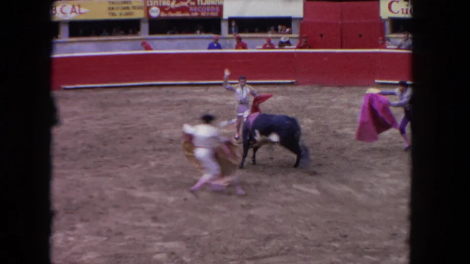 1962 Los Angeles 8mm frame showing three bullfighters in light costumes working around a black bull in a red arena with spectators above.
