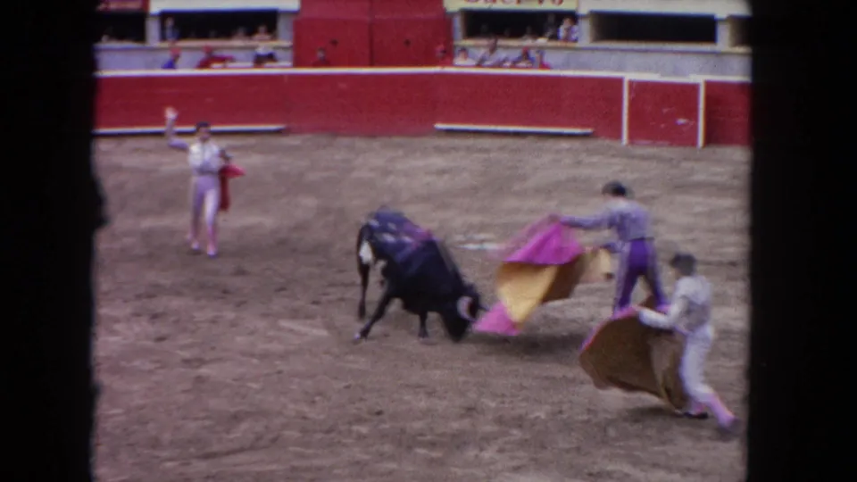 1962 Los Angeles arena frame with a black bull charging low toward a bullfighter's purple and yellow cape while other performers stand nearby.