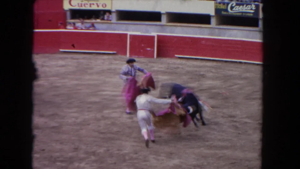 1962 Los Angeles 8mm frame showing bullfighters close to a black bull, with pink and yellow capes visible against the dirt floor and red barrier.