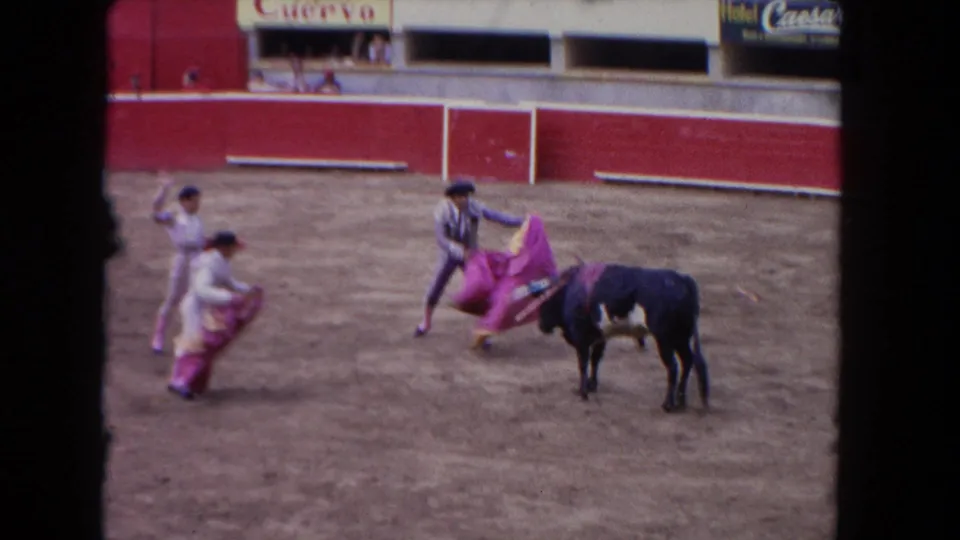1962 Los Angeles frame showing a black bull paused near two bullfighters with bright capes, with a third performer and arena signage in the background.