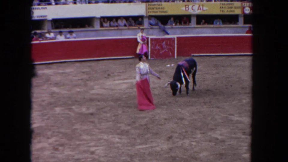 A matador pauses with the cape as a black bull lowers its head, with a woman standing by the arena wall in the 1962 Los Angeles footage.
