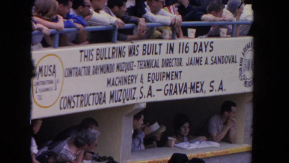 1962 8mm frame of a bullring construction sign and seated spectators, with Spanish-language text and arena advertising visible.