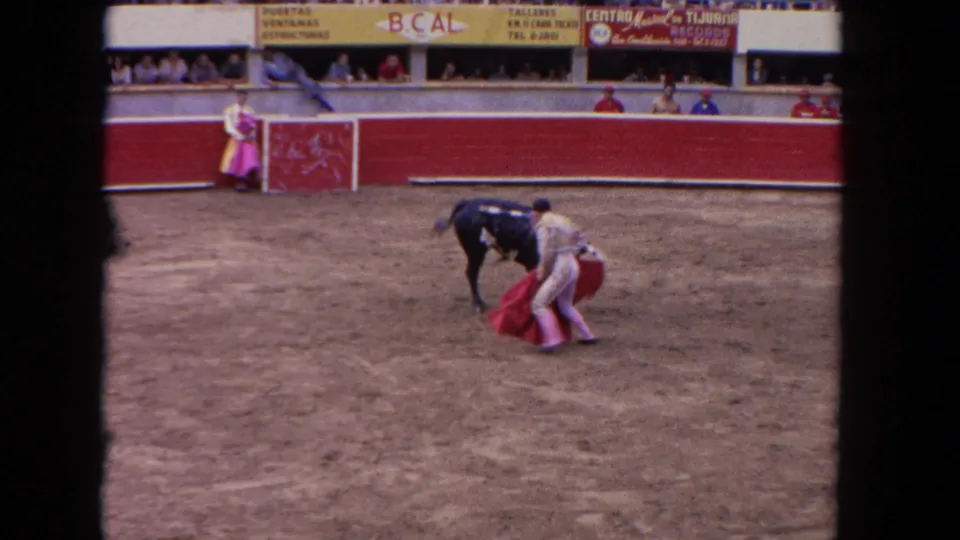 1962 8mm frame showing a matador using a red cape close to a black bull on sandy ground, with arena seating and barriers behind.