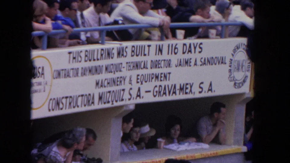 Spectators in 1960s clothing lean over blue railings above a Los Angeles bullring construction banner.