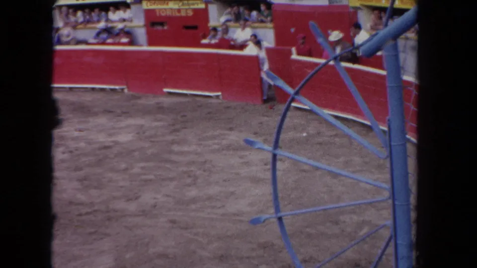 1962 Los Angeles bullring scene with red barriers, a dirt arena, distant spectators, and blue metal structure partly blocking the view.