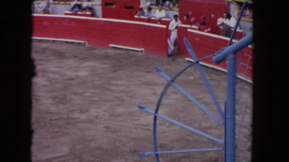 A man in white stands near the red wall of a 1962 Los Angeles bullring while spectators watch from behind the barrier.