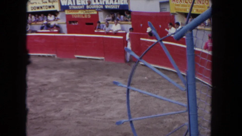 A dark bull runs near red barriers in a 1962 Los Angeles arena, with spectators and advertising signs behind it.