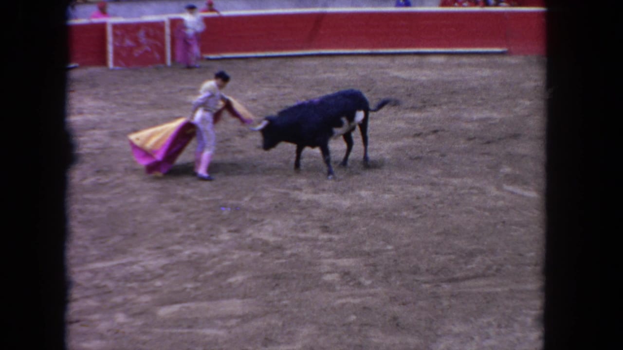 Matador holds a yellow and pink cape near a black bull in a dusty arena.