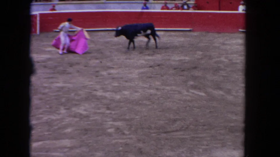 1962 Los Angeles 8mm frame showing a matador presenting a pink cape to a black bull in a red-walled arena with spectators visible above.