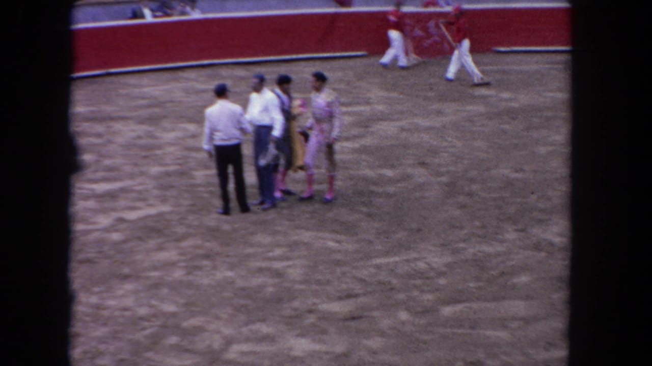 Two costumed matadors and two men in white shirts stand on a dirt arena floor near a red barrier.