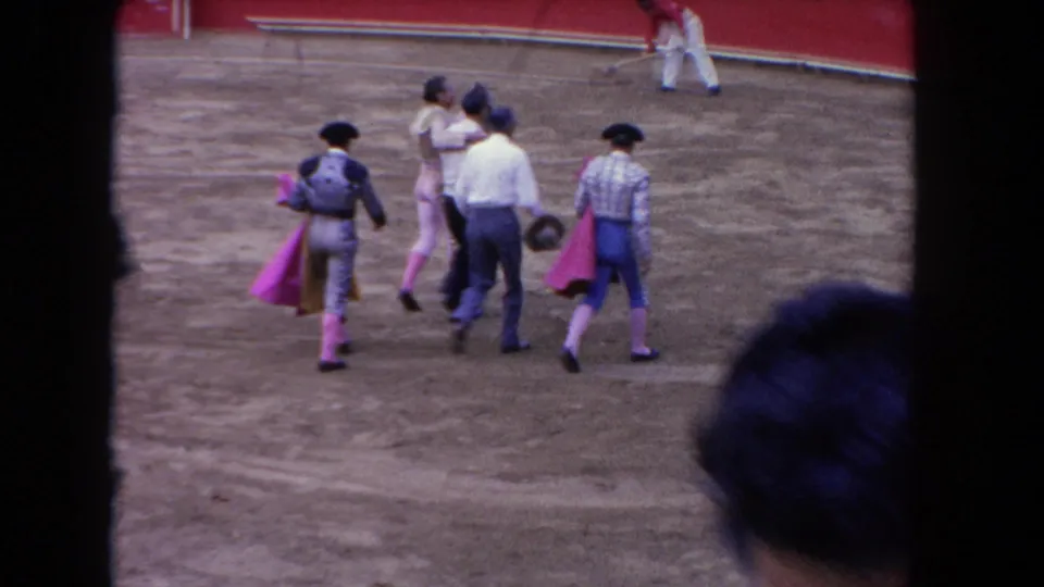 1962 Los Angeles home-movie frame showing matadors with capes and attendants crossing a sandy bullring from a spectator viewpoint.