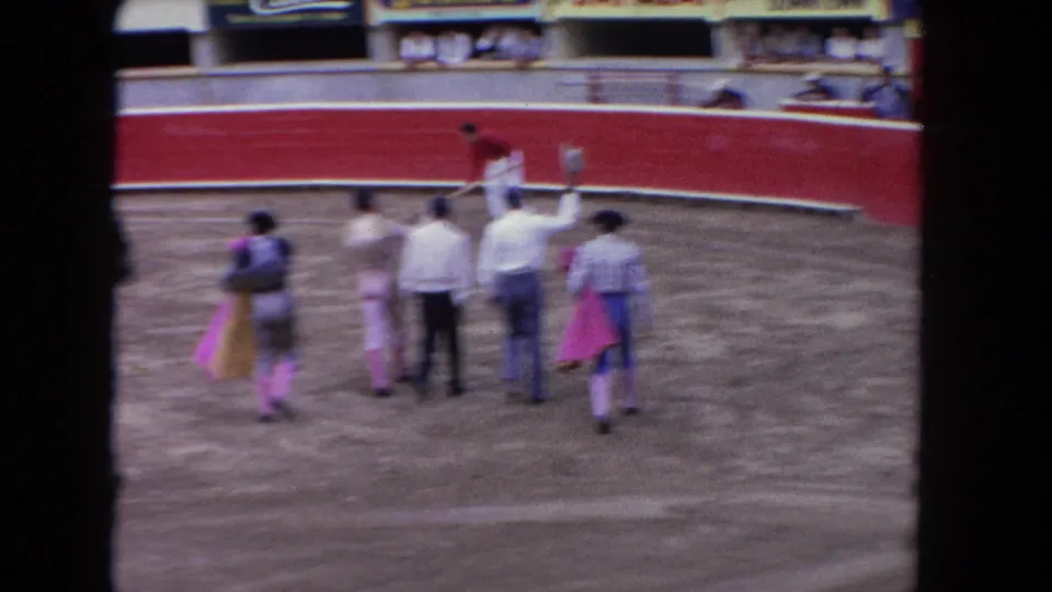 1962 Los Angeles arena view with matadors, attendants, spectators, and a worker raking the sandy ring near the red barrier.