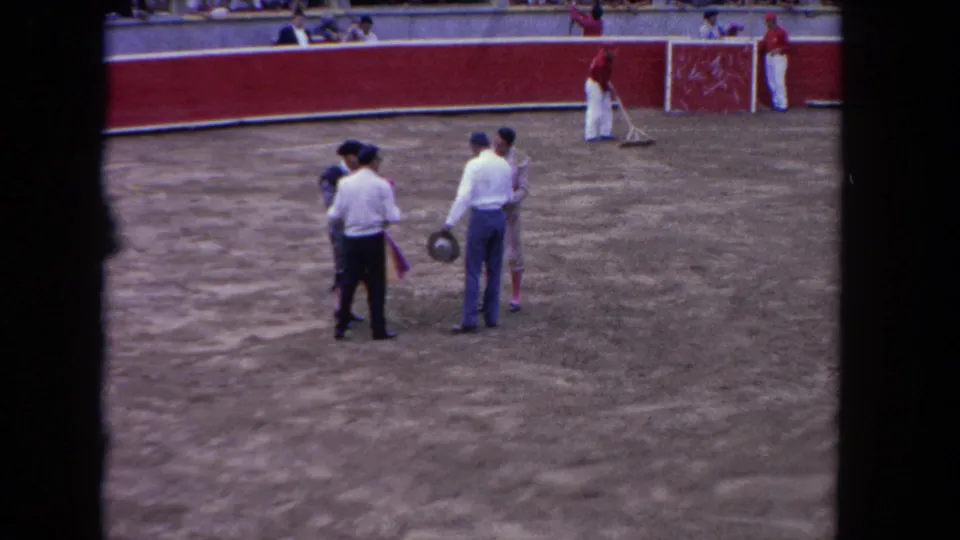 Home-movie frame from the Los Angeles bullfighting event showing matadors and attendants paused in the ring with capes visible.