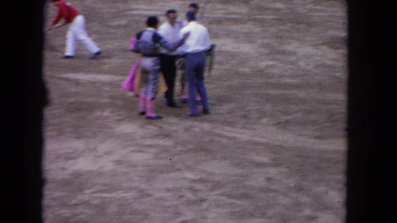 1962 Los Angeles bullring frame with casually dressed men and matadors standing together on the sandy arena floor.