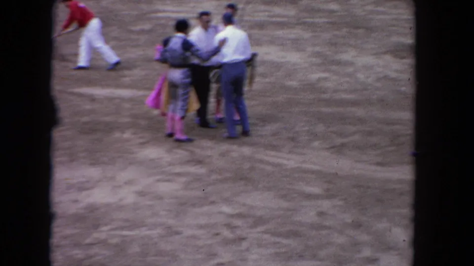 1962 Los Angeles bullring frame with casually dressed men and matadors standing together on the sandy arena floor.