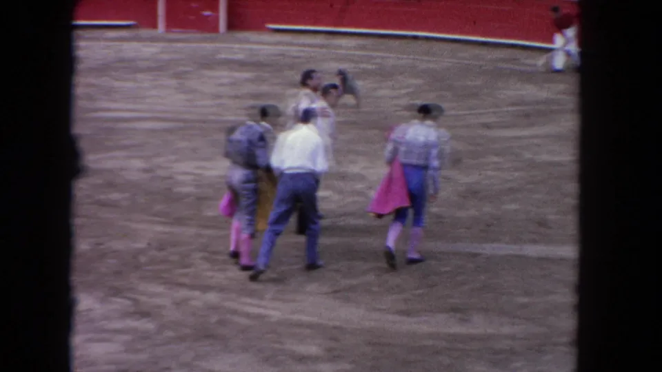 1962 Los Angeles bullring frame with men and matadors walking across the sandy arena near a red barrier wall.