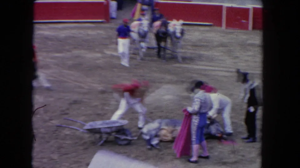 1962 Los Angeles 8mm frame with motion blur as attendants move beside a fallen bull, a wheelbarrow, red barriers, and horses in the ring.