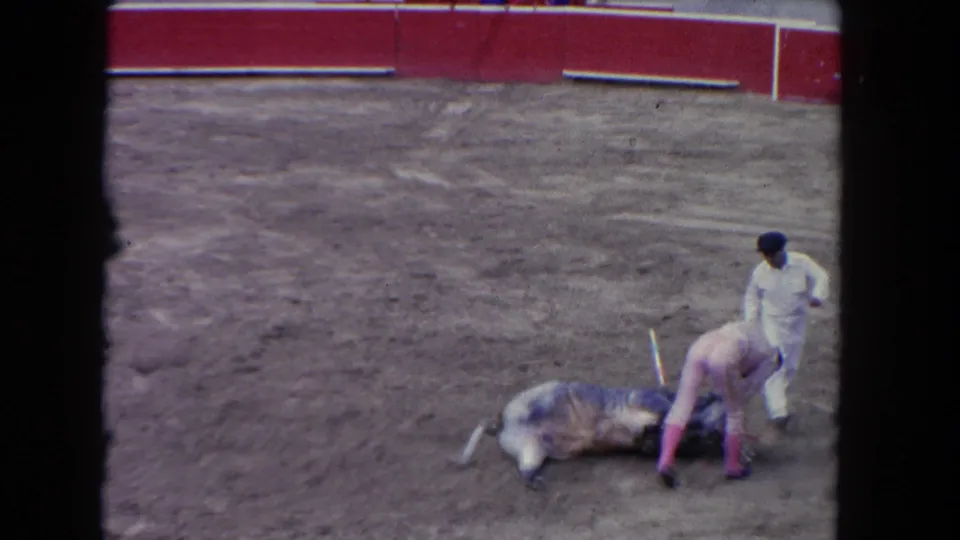 1962 Los Angeles home-movie frame showing a matador near a fallen bull in a dirt arena, with red barrier boards behind them.