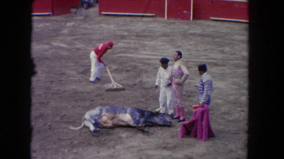 1962 Los Angeles home-movie frame with matadors, a cape holder, and a ring worker around a fallen bull in the arena.