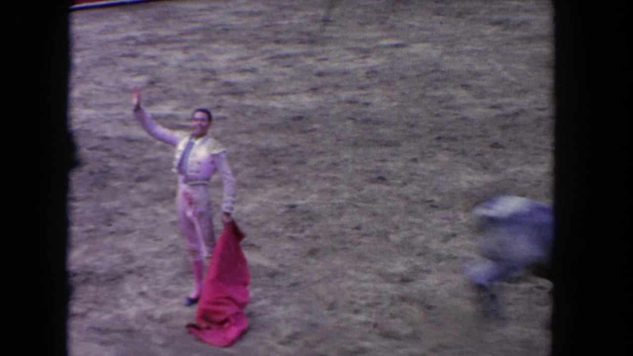1962 Los Angeles home-movie frame showing a matador with a red cape as a bull charges past on a dusty arena floor.