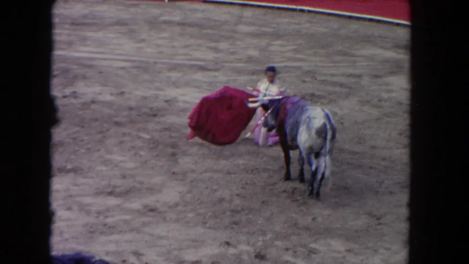 In 1962 Los Angeles home-movie footage, a matador in a pale costume holds a red cape beside a horned gray bull in a dusty ring.