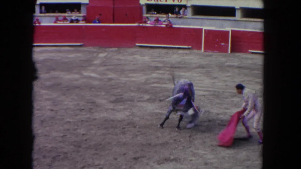 1962 Los Angeles home-movie frame showing the bull pivoting as the matador draws the red cape across the dirt arena.