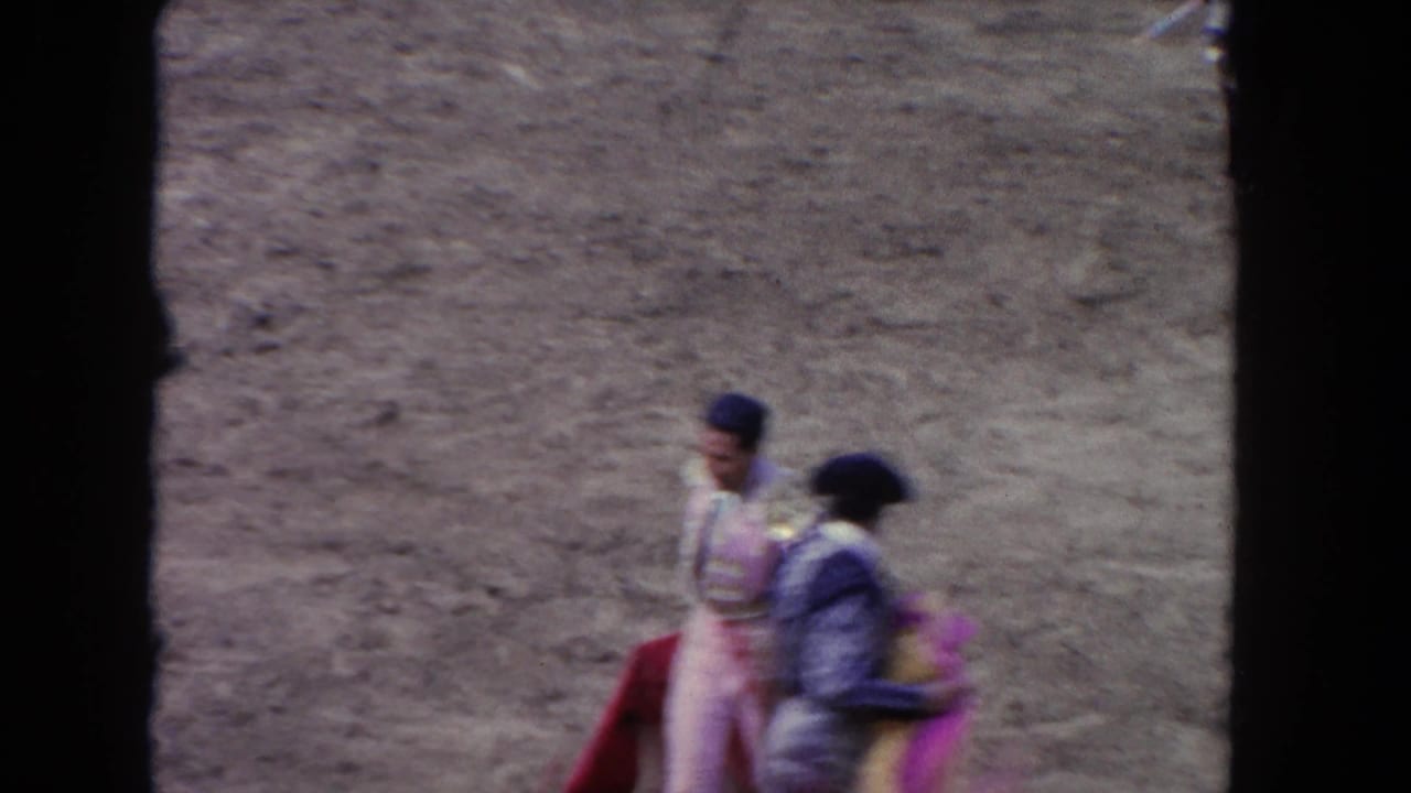 Matador and another person walk near a red arena barrier on a dusty bullring floor.