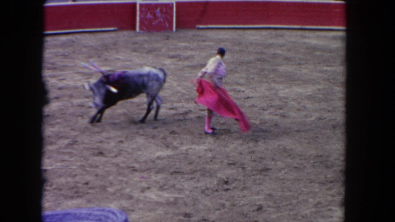 Matador holds a red cape as a bull approaches across a dirt arena.
