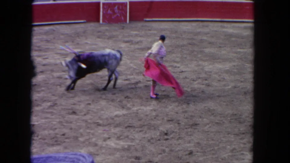 1962 Los Angeles 8mm frame showing a matador in white and pink facing a bull with a red cape in a dirt arena.