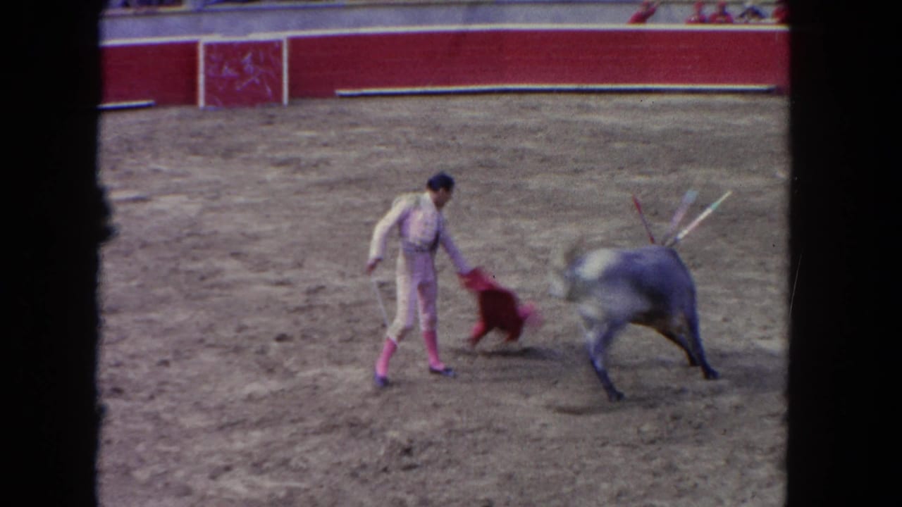 1962 Los Angeles home-movie frame showing a matador presenting a red capote to a grey bull in a sandy arena.